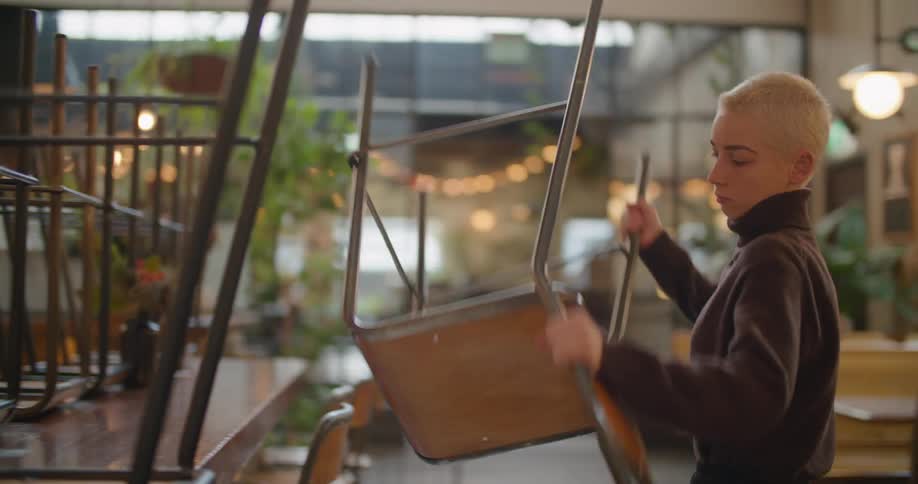 A worker in a restaurant pulling the chairs off from on top of a table and fixing them in a seating position