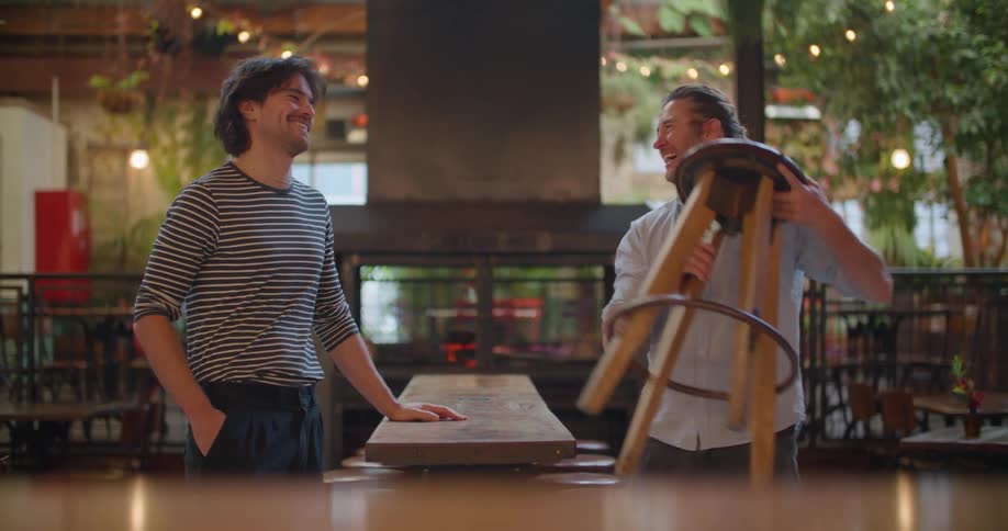 Two men in a restaurant pulling stools down from the top of a table while laughing and talking to each other