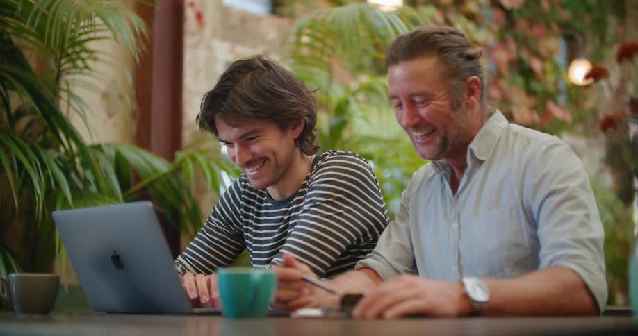 Two men sit in a restaurant and laugh and look at a laptop