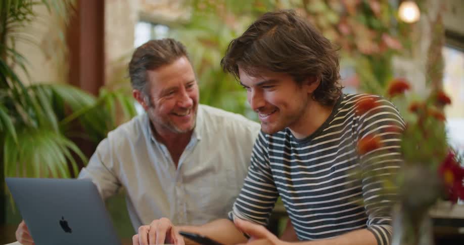 Two men smiling and talking while using a laptop over a desk in a room full of plants