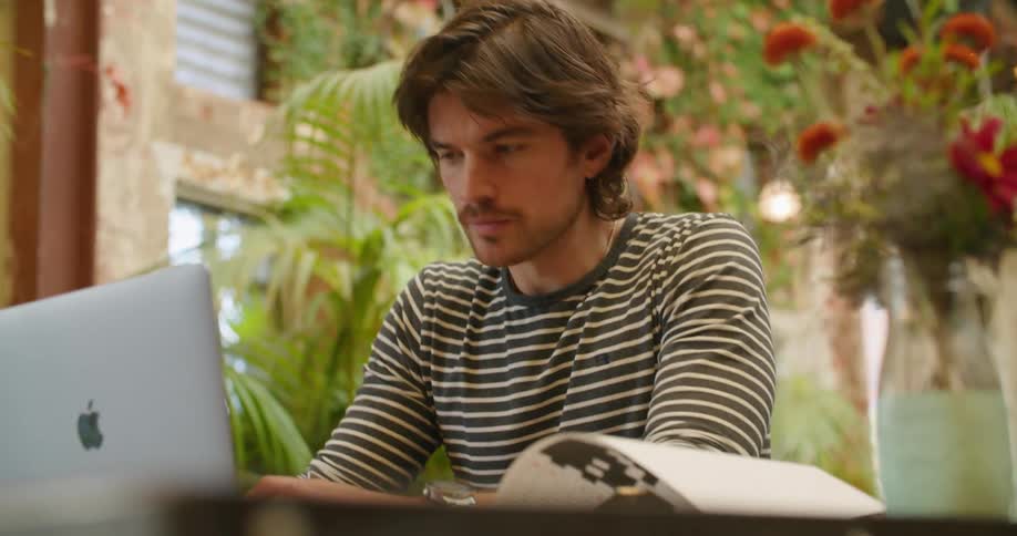 A man sits outside working on a laptop and looking at a notebook
