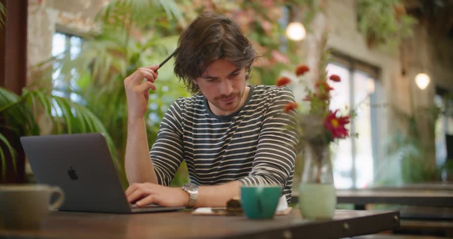 A man sits at an outside table working on a laptop and scratching his head with a pen in confusion