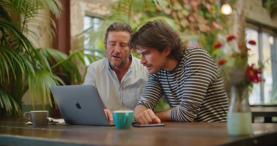 Two men sit together at an outside table looking at a laptop