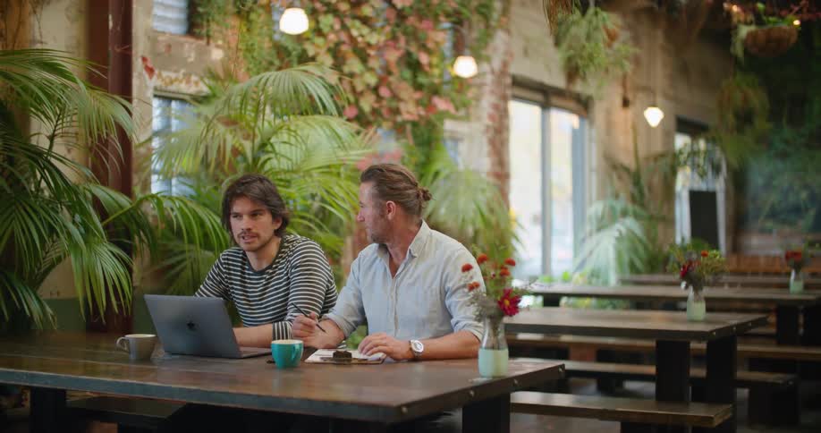 Two men sit together at a table outside with a laptop