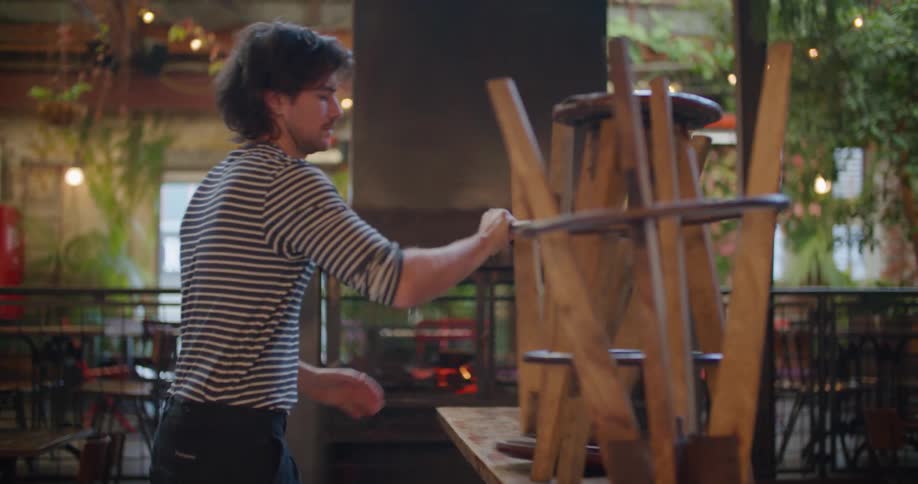 A man working in a restaurant takes stools off of tables and sets them on the ground