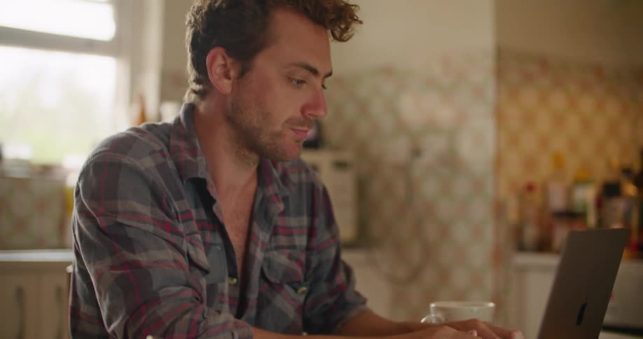 A man sitting on a chair at the kitchen table typing on a laptop