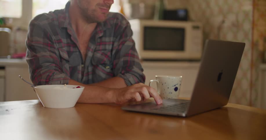 A man sits at a kitchen table with a laptop and takes a drink from a mug