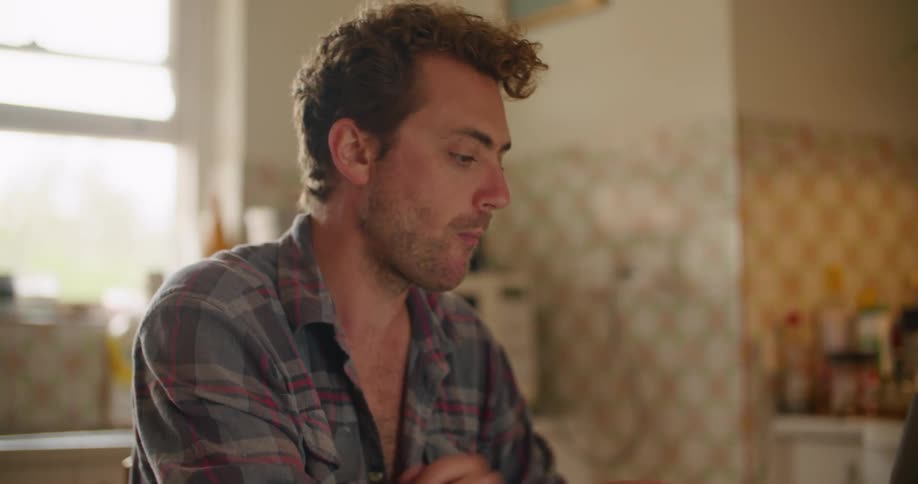Man with a plaid shirt sitting at table drinks coffee and works on his laptop in the kitchen
