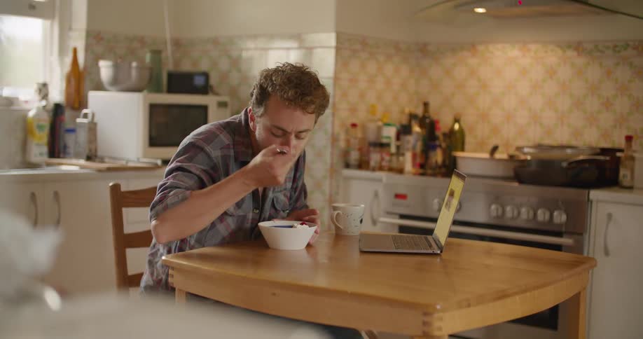 A man sitting on a chair at a table in a kitchen and using a laptop while eating