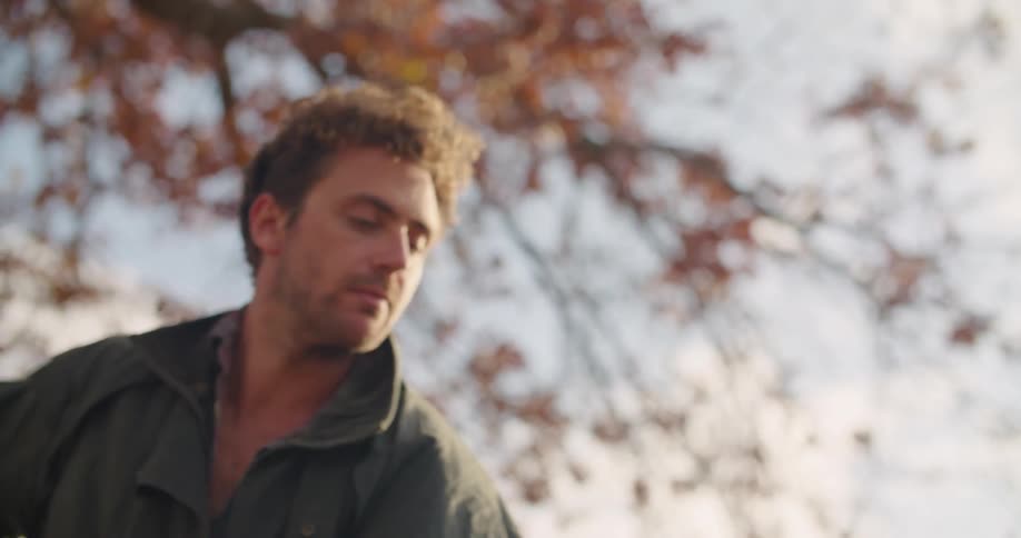 Outdoors in front of light blue sky and tree with orange leaves a man focuses intently on the ground while taking some action coordinated arm movements