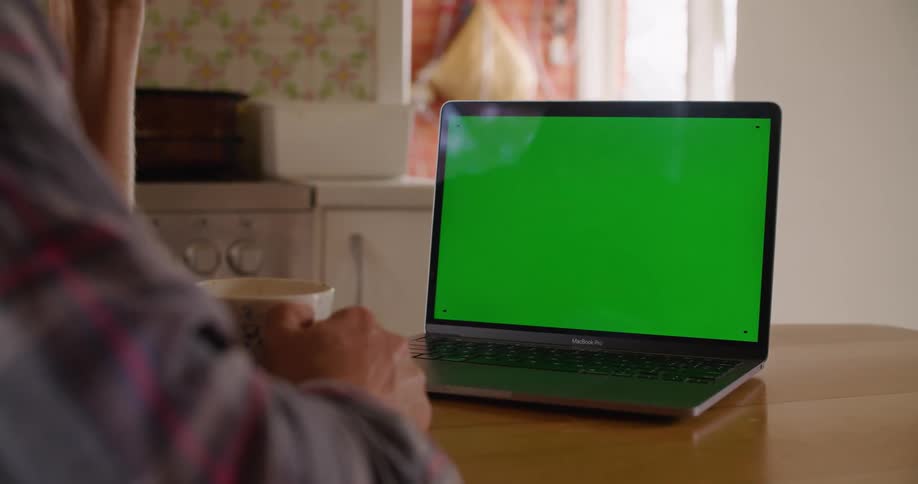 Man sitting at table holds a cup and looks at a laptop with green screen in the kitchen