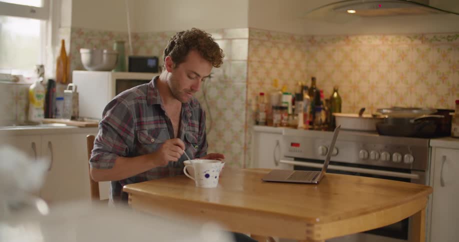 Man with a shirt sitting at table eats breakfast and looks at laptop in the kitchen