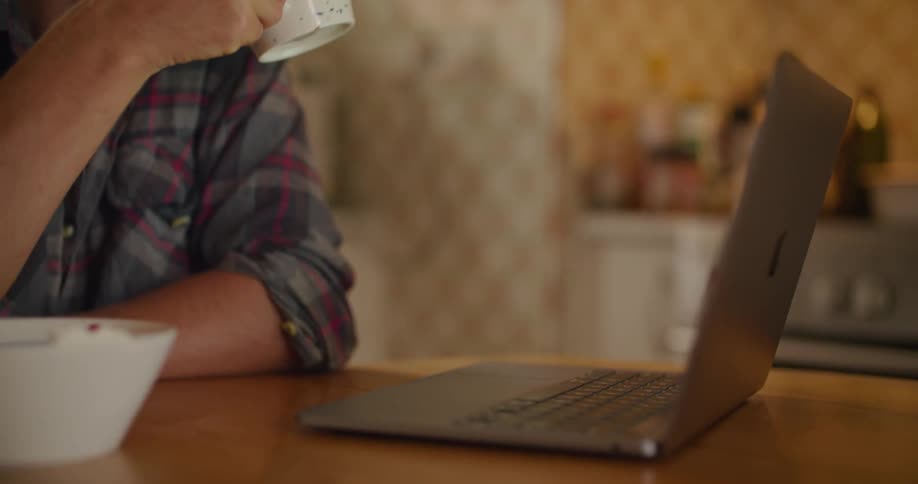 A man sits at a small home table looking at a laptop and drinks from a mug
