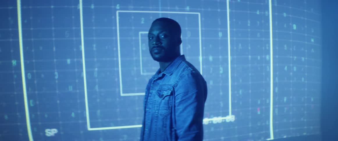 A man with a jacket in a room standing and staring with a screen in the background