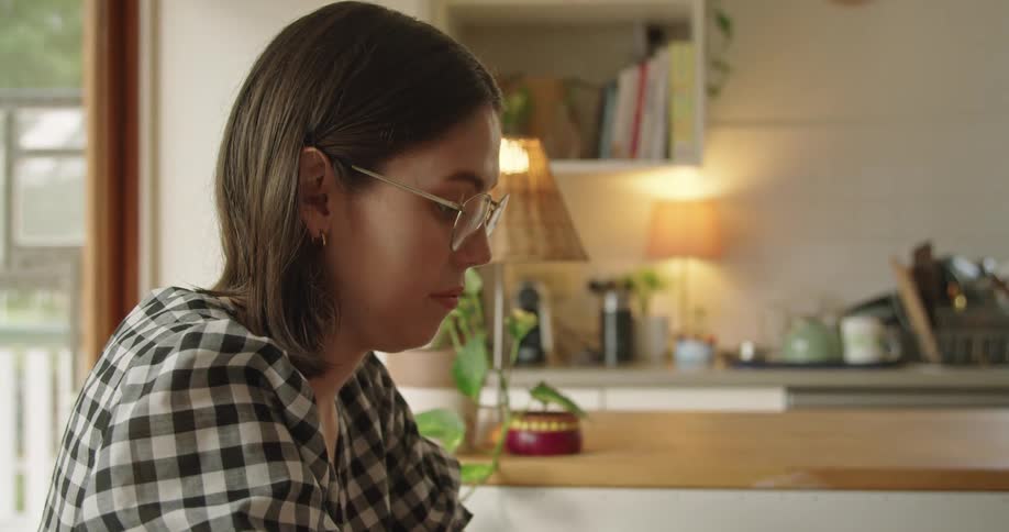 Woman with glasses and a plaid shirt sits at table works on laptop in the kitchen