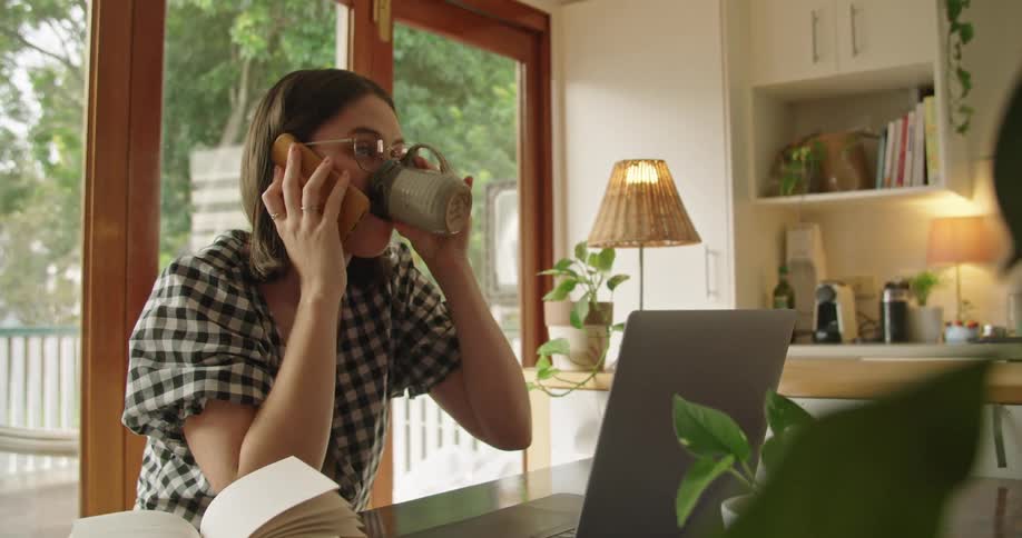 A woman sits at a table talking on the phone and takes a drink from a mug
