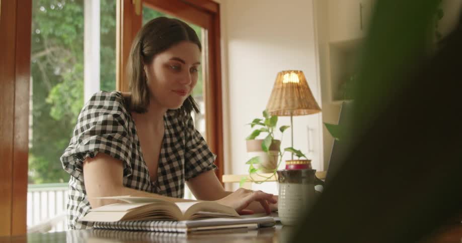 A woman in a room sitting at a table typing on a laptop with a lamp and a door in the background
