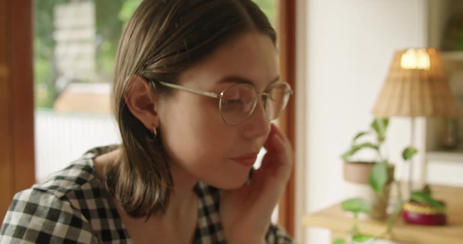 Woman sitting at table cleans her glasses and uses her laptop in the kitchen