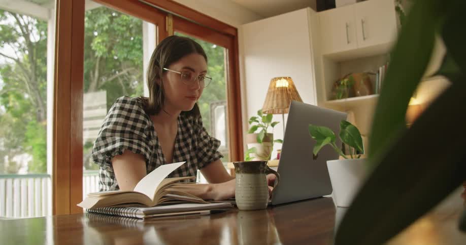 Woman with glasses and a plaid shirt sits at table and works on laptop in the kitchen at home