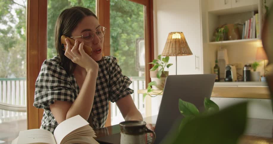 Woman working from home talks on the phone while using a laptop