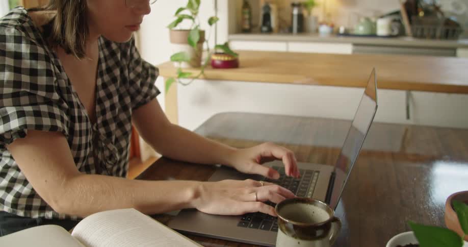 Woman with a blouse and glasses looks at a book and types on a grey laptop in the kitchen