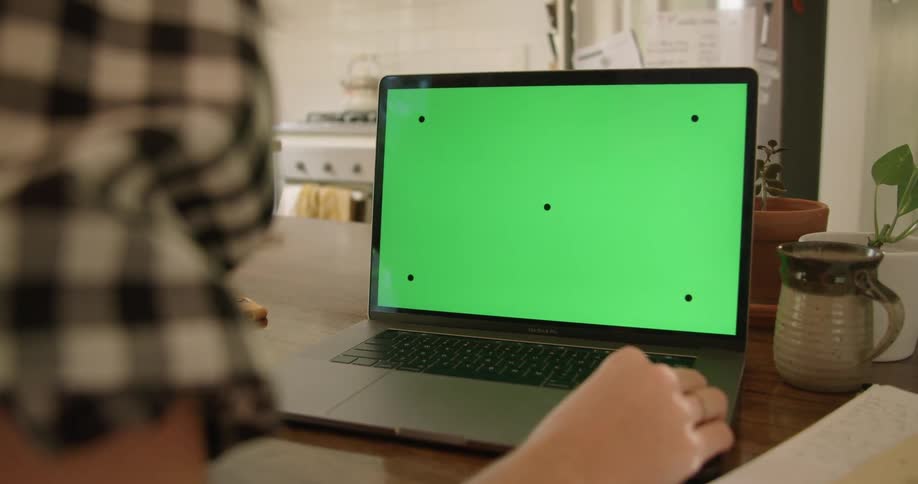 A married person in a checkered shirt sits at a table facing a brightly lit kitchen and rests their right hand on a macbook pro displaying a green screen