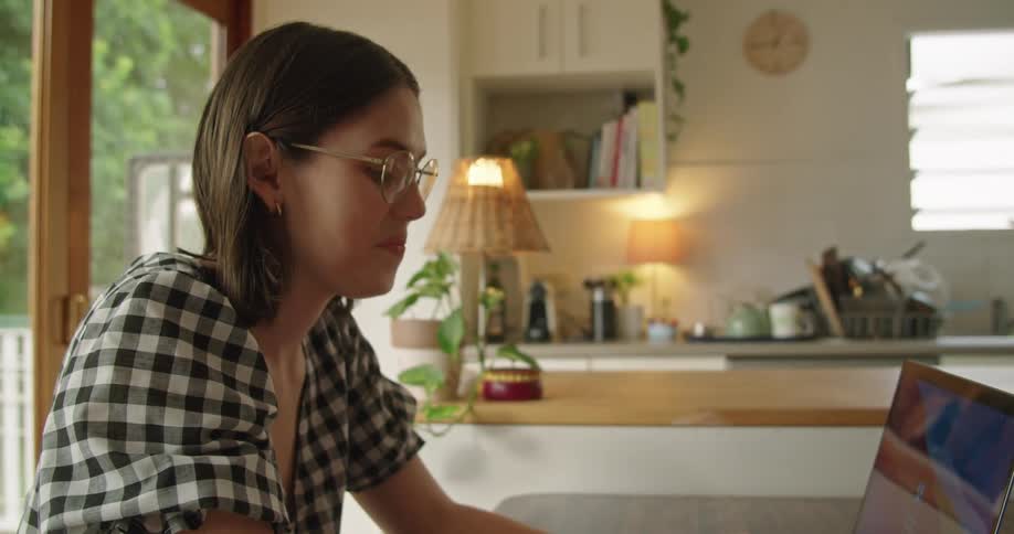 Woman with glasses and a plaid shirt sits at table and works on laptop smilingly in the kitchen