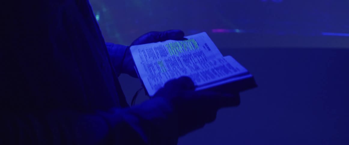 A man flips through a small book while standing in front of a projector screen showing a video game