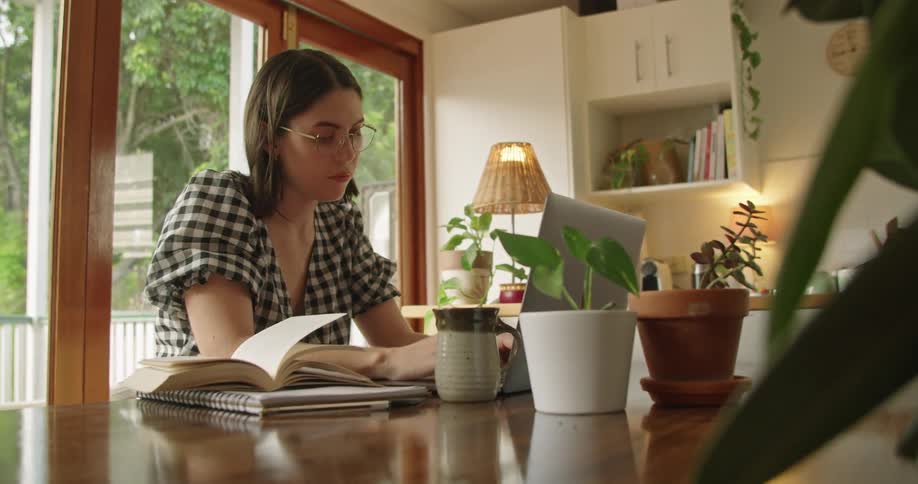 Woman with a blouse glasses sitting at table uses a grey laptop in the kitchen