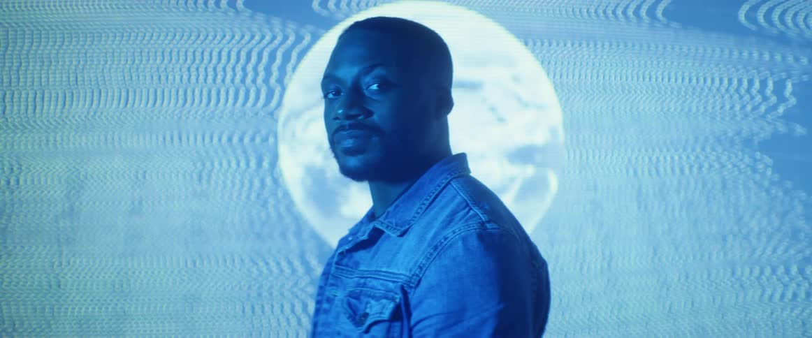 A man standing in a room with a blue hue in front of a digital screen with a spiraling planet earth staring at the camera
