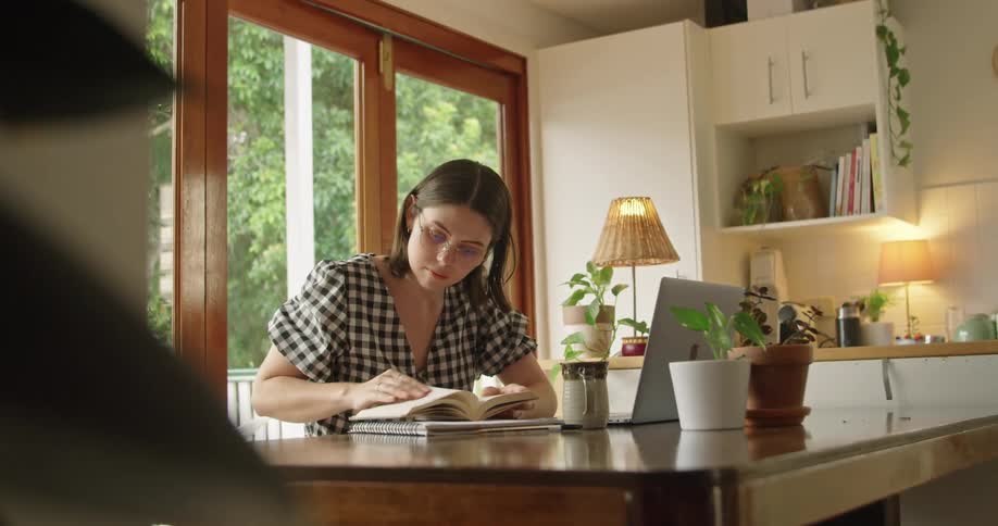 A woman flipping through a book and typing on a laptop while sitting in a home office