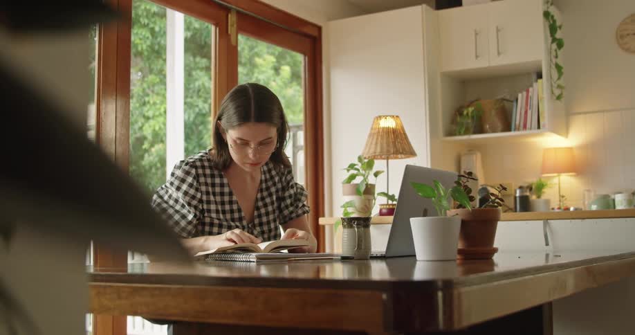 A woman sitting at a table looking through a book and typing on a laptop computer