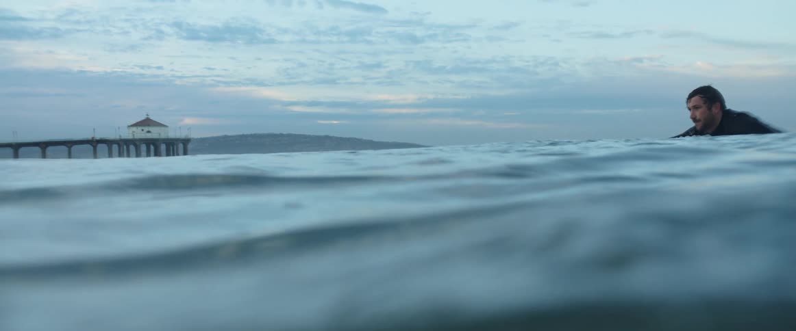 Man in a diving suit swims with a surfboard with a pier in the background at dusk