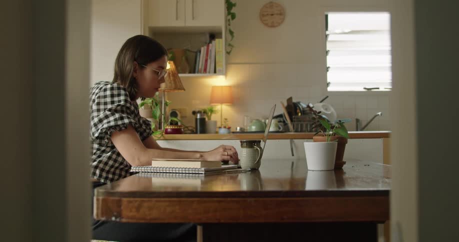 A woman sitting at a kitchen table looking at a smart phone and then typing on a laptop