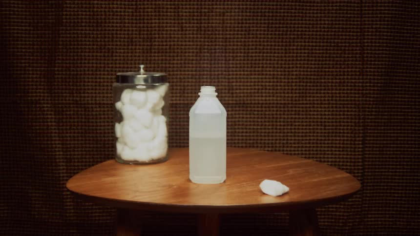 A person places a bottle of rubbing alcohol on a table beside a jar of cotton balls