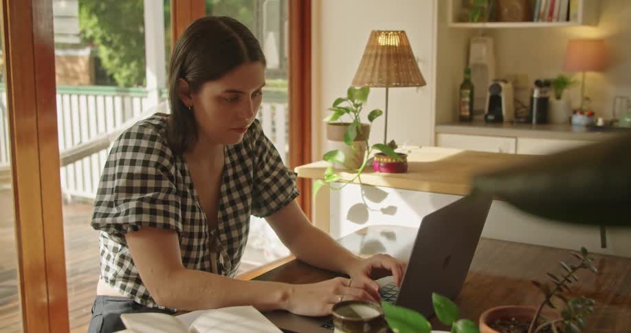 A woman typing on a laptop computer inside of a room sitting at a table next to a window during the day