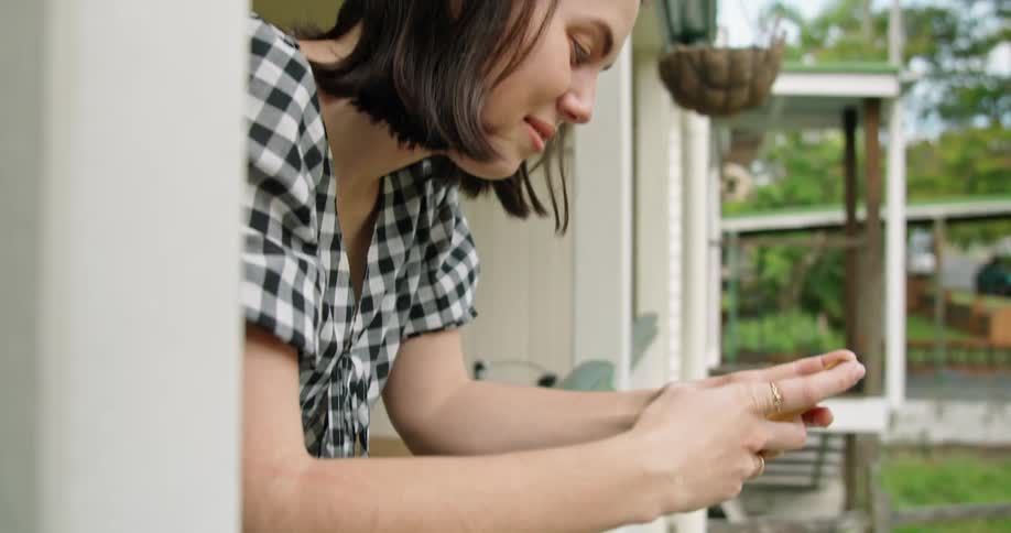 A woman leaning on a banister and using a smart phone with trees the sky and clouds in the background
