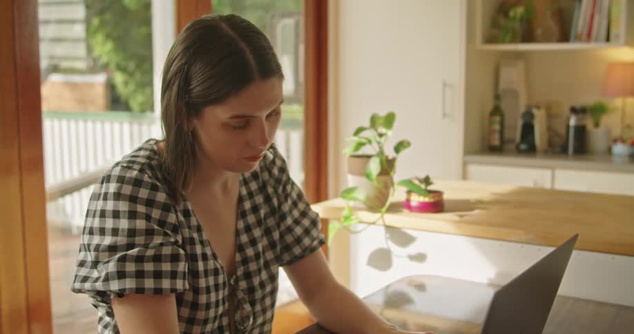Woman with a blouse sitting at table works on grey laptop in the kitchen