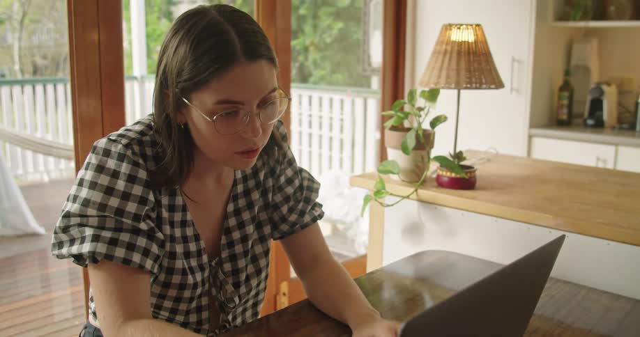 Woman with glasses and a plaid shirt sits at table looks at a book and works on a laptop in the kitchen with the terrace in the background