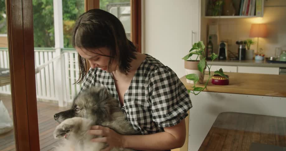 Woman with a blouse sitting at table strokes the grey dog in the kitchen at home