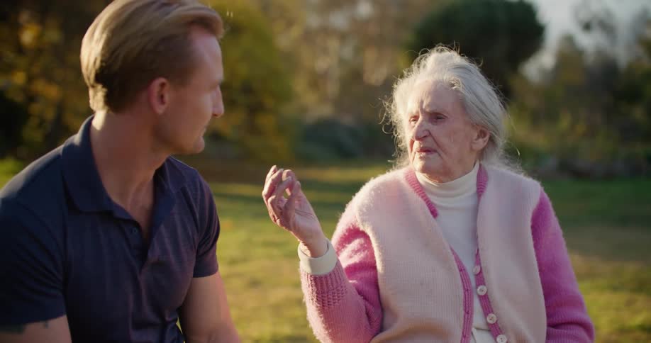 Man watches and listens to elderly woman talk and gesture while they sit outdoors