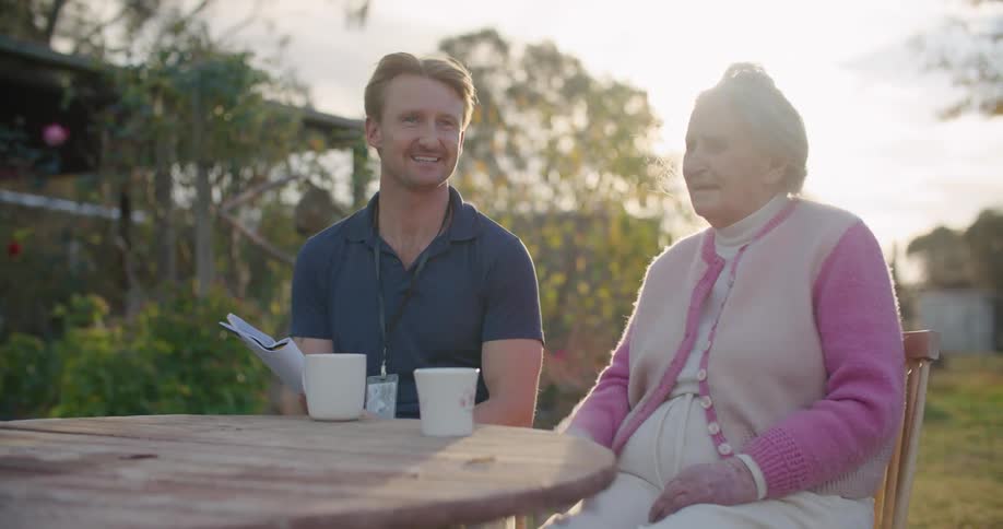 Man wearing badge on lanyard sits outdoors at wooden table talking with elderly woman