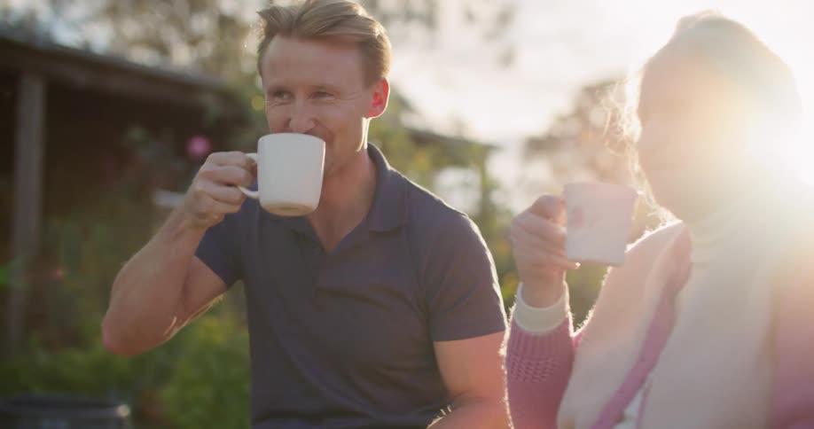 Man with a dark blue polo shirt and elderly woman with a white vest with a pink sweater raise white cups to drink with the sun going down in the background in the garden