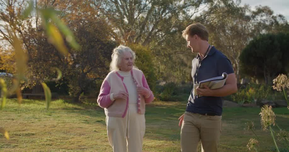 A man carrying an open notebook and wearing an id badge on a lanyard talks with an elderly woman in a garden