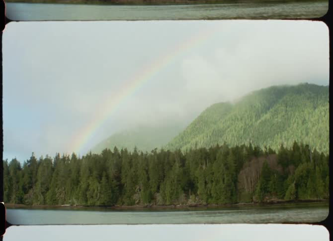 A rainbow in a foggy sky extending over a forest and mountains viewed from across a river