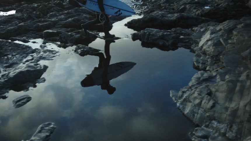 A man carries his surfboard on the rocks by the shore
