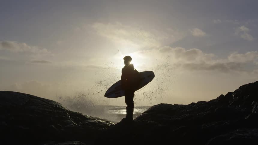 A male stands on the shore holding a surfboard as water sprays up