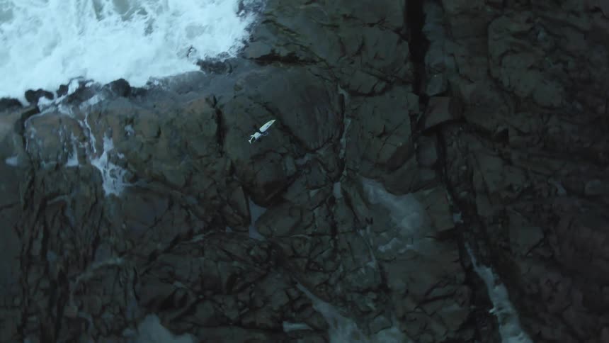 A person with a surfboard on a rocky shore with waves of water crashing