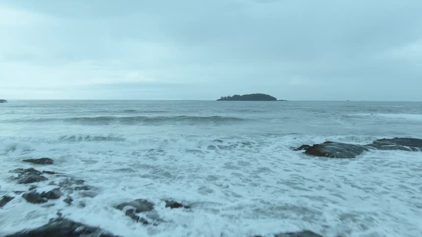 Person holding surfboard stands on rocky shore looking toward island on horizon under cloudy sky