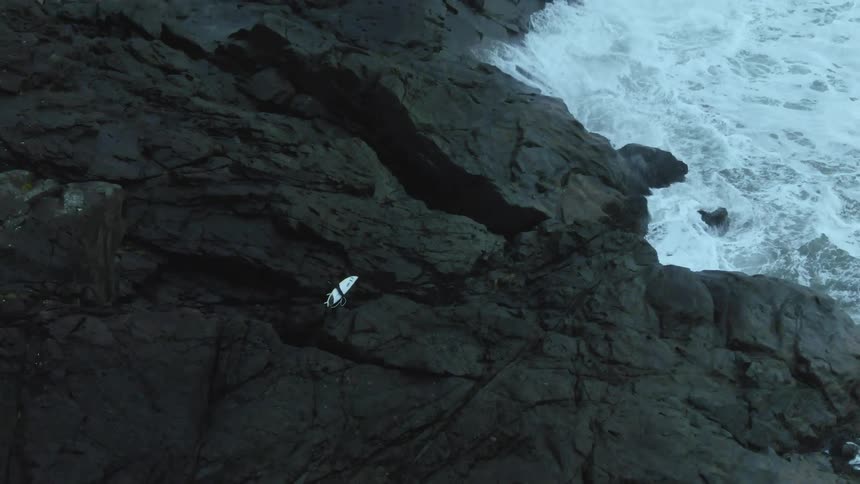 Young man holding a surfboard walks on a rock promontory in front of the coast